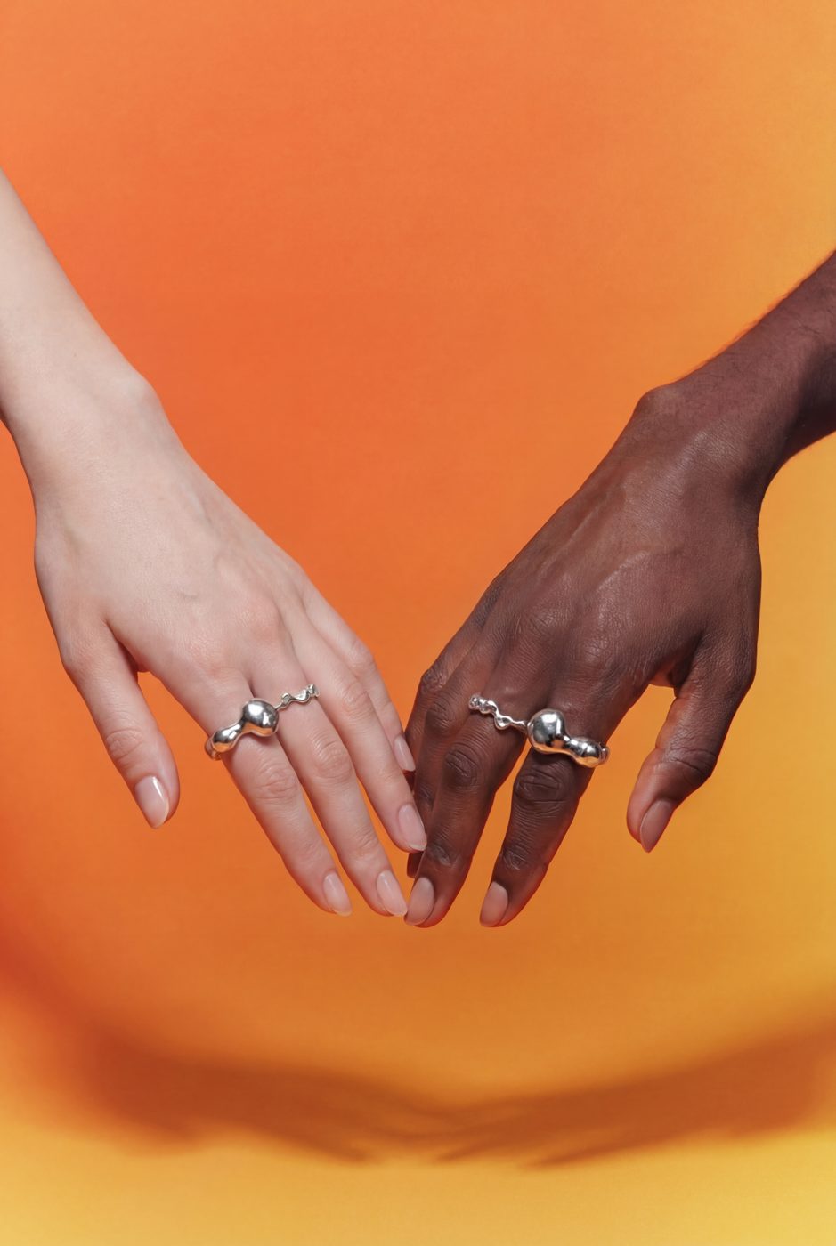 Two hands wearing polished silver rings on an orange background